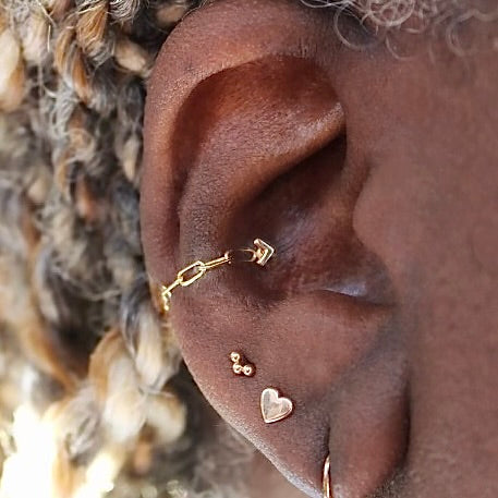 Photo of an Ahimsa Piercing Studio client with 4 piercings on her right ear. First lobe piercing showing part of a dangling earring, second lobe piercing featuring BVLA's "Flat Heart", third lobe piercing featuring BVLA's "Tri Bead Arc" and a conch piercing fearuting BVLA's "Flat V" with a link chain attached to the front and back. Mix of yellow and rose gold