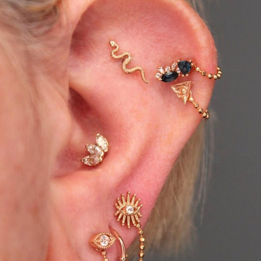 Photo of an Ahimsa Piercing Studio client with 8 piercings on her left ear ear. Conch, and 3 visible lobe piercings in rose gold with CZ and 3 stacked helix piercings with jewelry in rose gold with London blue topaz and 2 beaded chains and a flat piercing featuring a snake rose gold