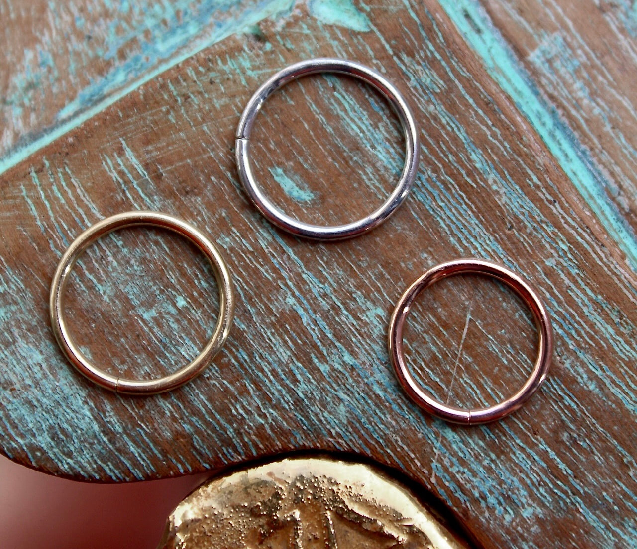 BVLA's "Seam Ring" shown from left to right in 14k Yellow gold, 14k White gold and 14k Rose gold. shown on a background featuring a piece of wood with blue paint on top and a small golden piece on the bottom of the photo