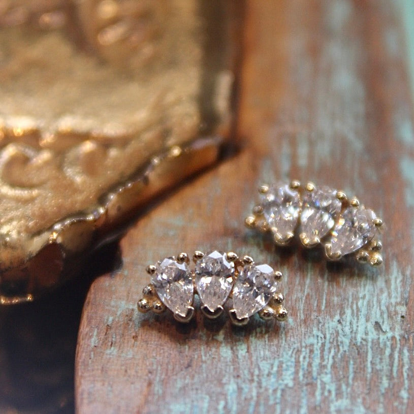 BVLA's "Pear Panaraya" shown twice in 14k Yellow gold with CZ. Shown on a background featuring a piece of wood with blue paint on bottom right and a small golden piece on the top left of the photo