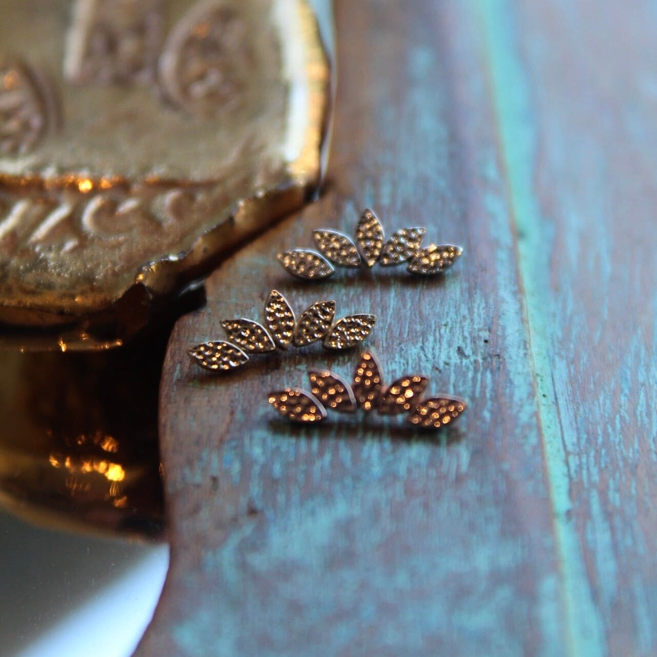 BVLA's "Hammered Serenity" shown from top to bottom in 14k Yellow gold, 14k White gold and 14k Rose gold. Shown on a background featuring a piece of wood with blue paint on bottom and a small golden piece on the top left of the photo