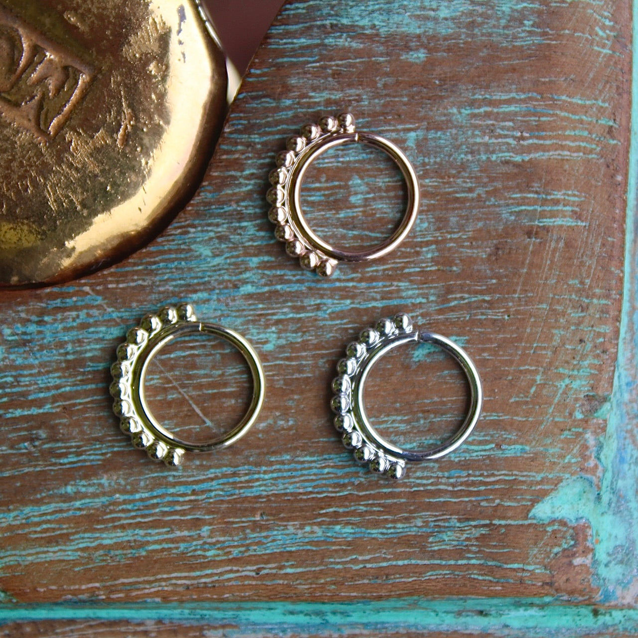 BVLA's "Latchmi Seam Ring" shown from left to right in 14k Yellow gold, 14k White gold and 14k Rose gold on top. Shown on a background featuring a piece of wood with blue paint on bottom and a small golden piece on the top left of the photo