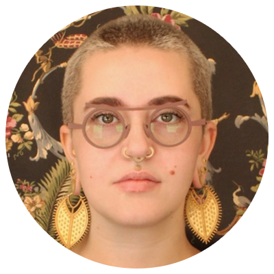 Ahimsa Piercing Studio staff member Tallulah wearing glasses and yellow gold facial jewelry and large yellow gold earrings standing in front of the studio's wallpaper