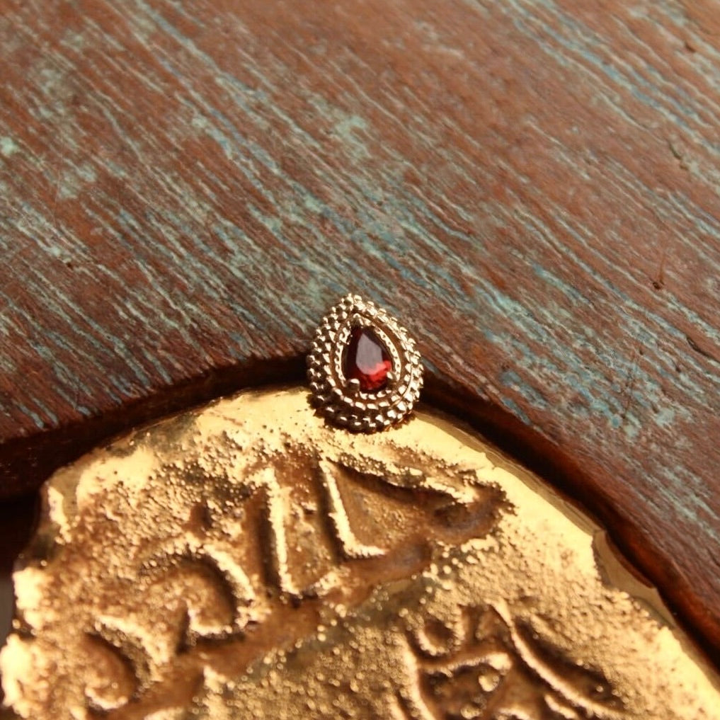 BVLA's "Afghan Pear" in 14k Yellow gold with Garnet shown on a background featuring a piece of wood with blue paint on top and a small golden piece on the bottom of the photo