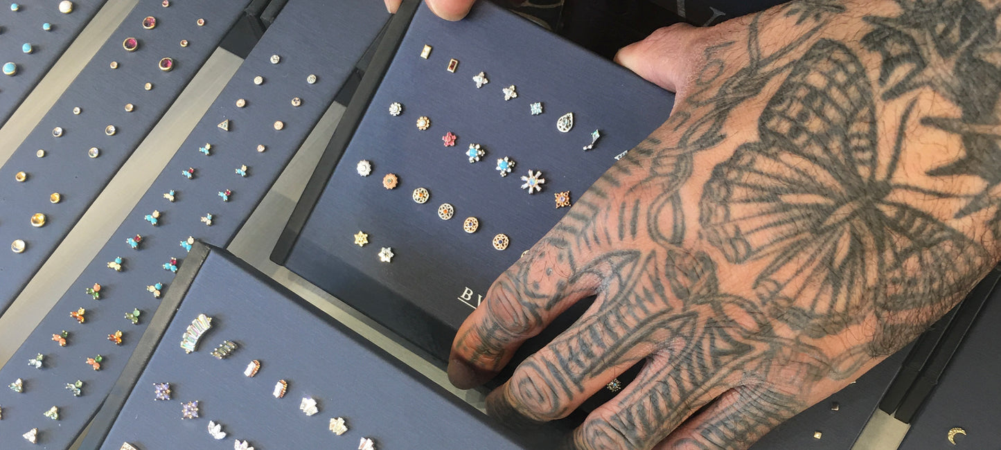 A tattooed hand adjusting a Navy Blue BVLA Jewelry display featuring an assortment of Yellow, rose and white gold jewelry with colored gemstones