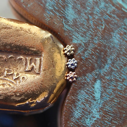 BVLA's "7 Bead Flower" shown from top to bottom in 14k Yellow gold, 14k White gold and 14k Rose gold. Shown on a background featuring a piece of wood with blue paint on the right and a small golden piece on the left of the photo