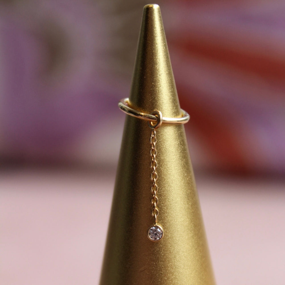 BVLA's "Bezel Dangle Charm" in 14k Yellow gold with a CZ Bezel at the bottom of a link style chain attached to a seam ring. Shown on a pointed cone gold colored jewelry display with the chain draping down.