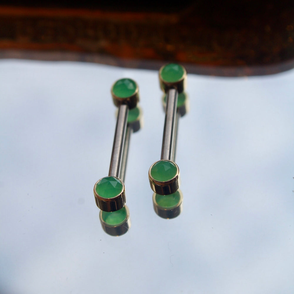 BVLA's "Bezel Side Pin" shown 4 times in 14k Yellow gold with chrysoprase, 2 each attached to 2 Anatometal "Straight Barbell" in titanium. Shown on a background featuring a mirror and a small golden piece on the top