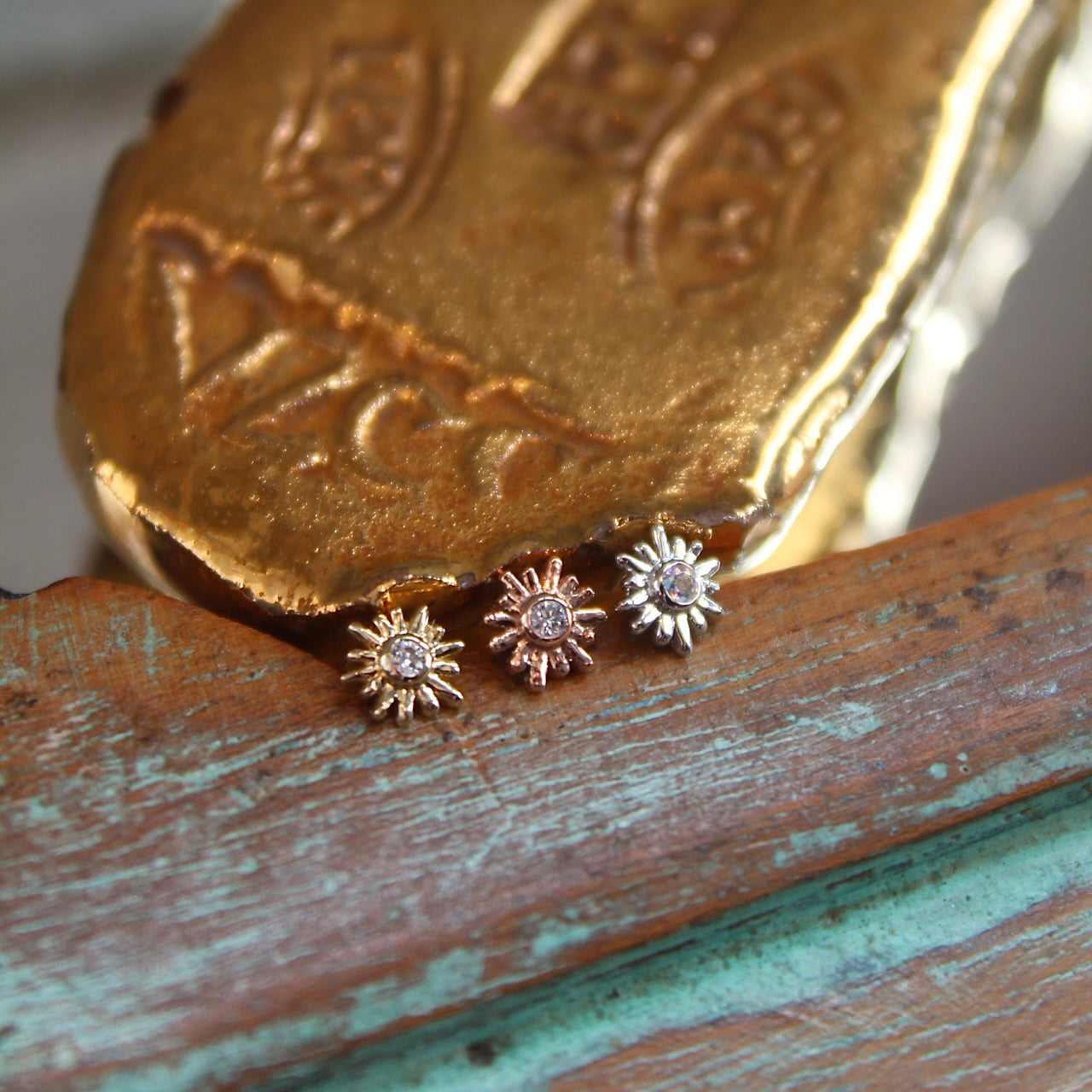 
                  
                    BVLA's "Sun Ray" shown from left to right in 14k Yellow gold with diamond, 14k Rose gold with Diamond and 14k White gold with diamond. Shown on a background featuring a piece of wood with blue paint on bottom and a small golden piece on the top of the photo
                  
                