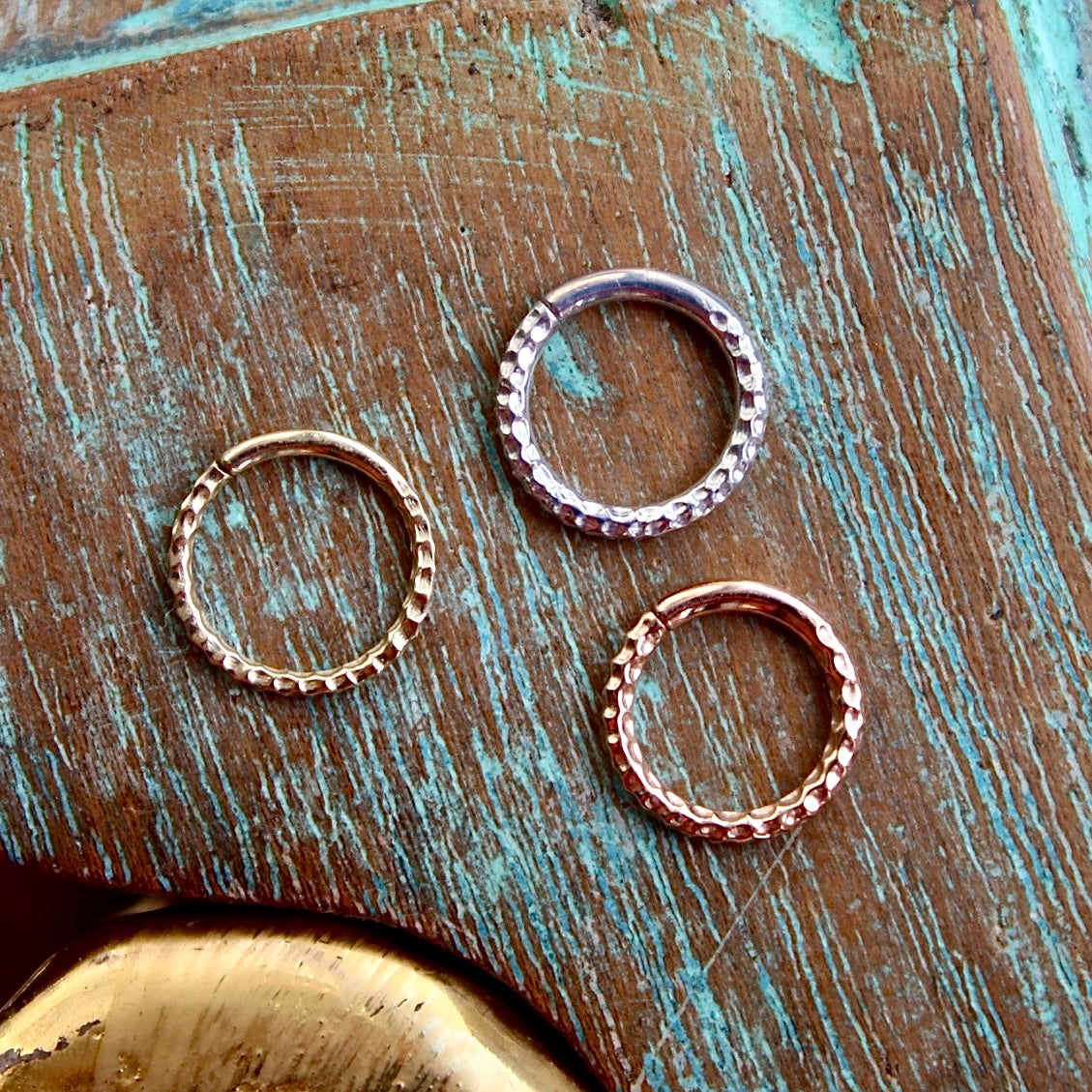 BVLA's "Hammered Seam Ring" shown from left to right in 14k Yellow gold, 14k White gold and 14k Rose gold. Shown on a background featuring a piece of wood with blue paint on top and a small golden piece on the bottom of the photo