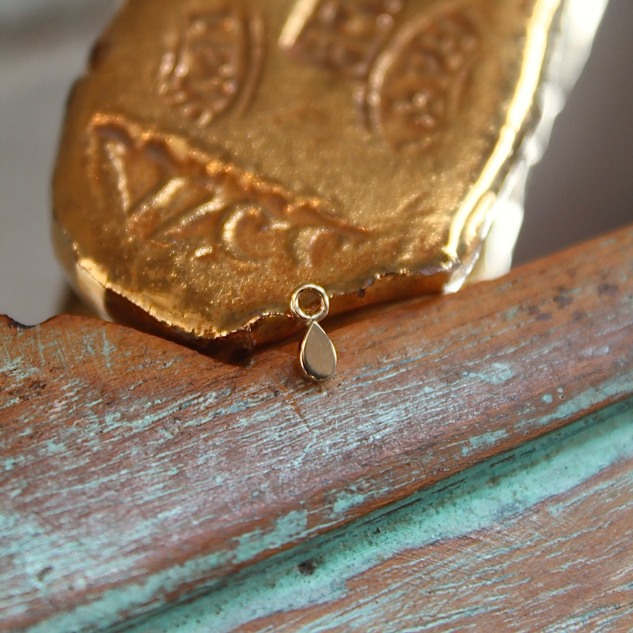 
                  
                    BVLA's "Flat Tear Charm" shown in 14k Yellow gold with the point up attached to a jump ring. Shown on a background featuring a piece of wood with blue paint on bottom and a small golden piece on the top of the photo
                  
                