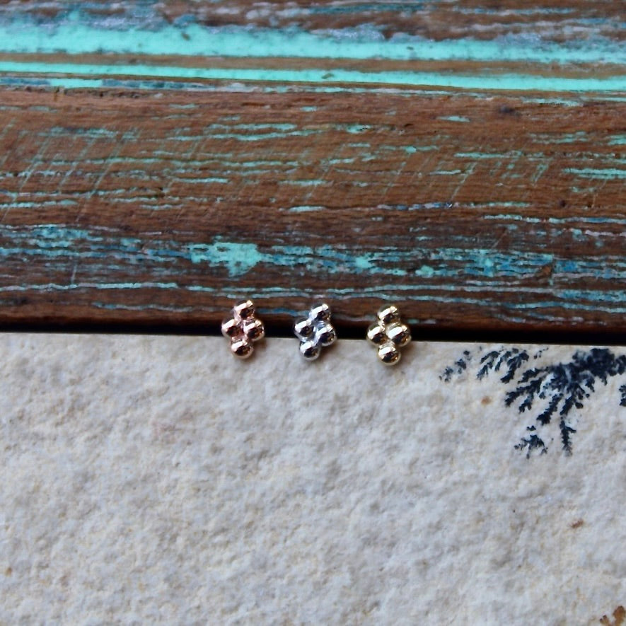 
                  
                    BVLA's "Quad Bead Cluster" shown from left to right in 14k Rose gold, 14k White gold and 14k Yellow gold. shown on a background featuring a piece of wood with blue paint on top and a light grey stone on the bottom of the photo
                  
                
