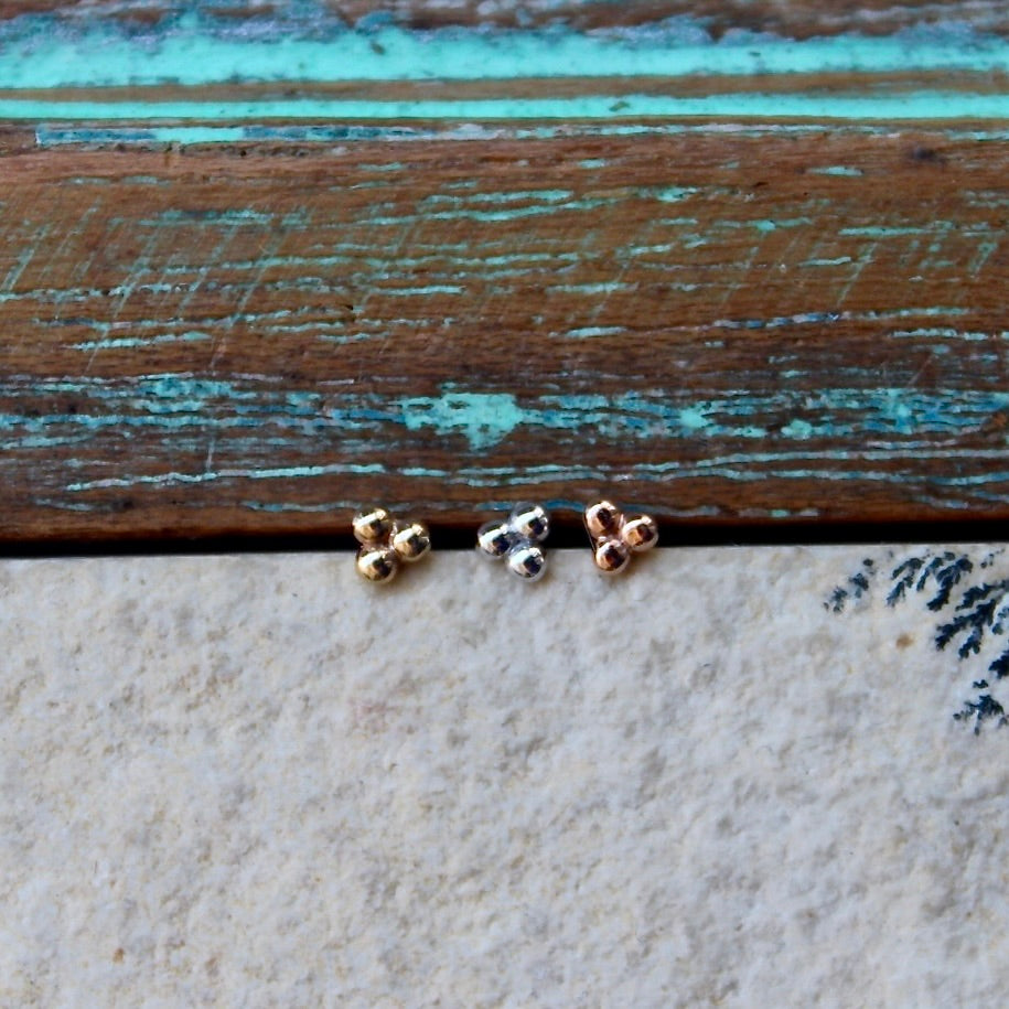 BVLA's "Tri Bead Cluster" shown from left to right in 14k Yellow gold, 14k White gold and 14k Rose gold. Shown on a background featuring a piece of wood with blue paint on top and a light grey stone on the bottom of the photo