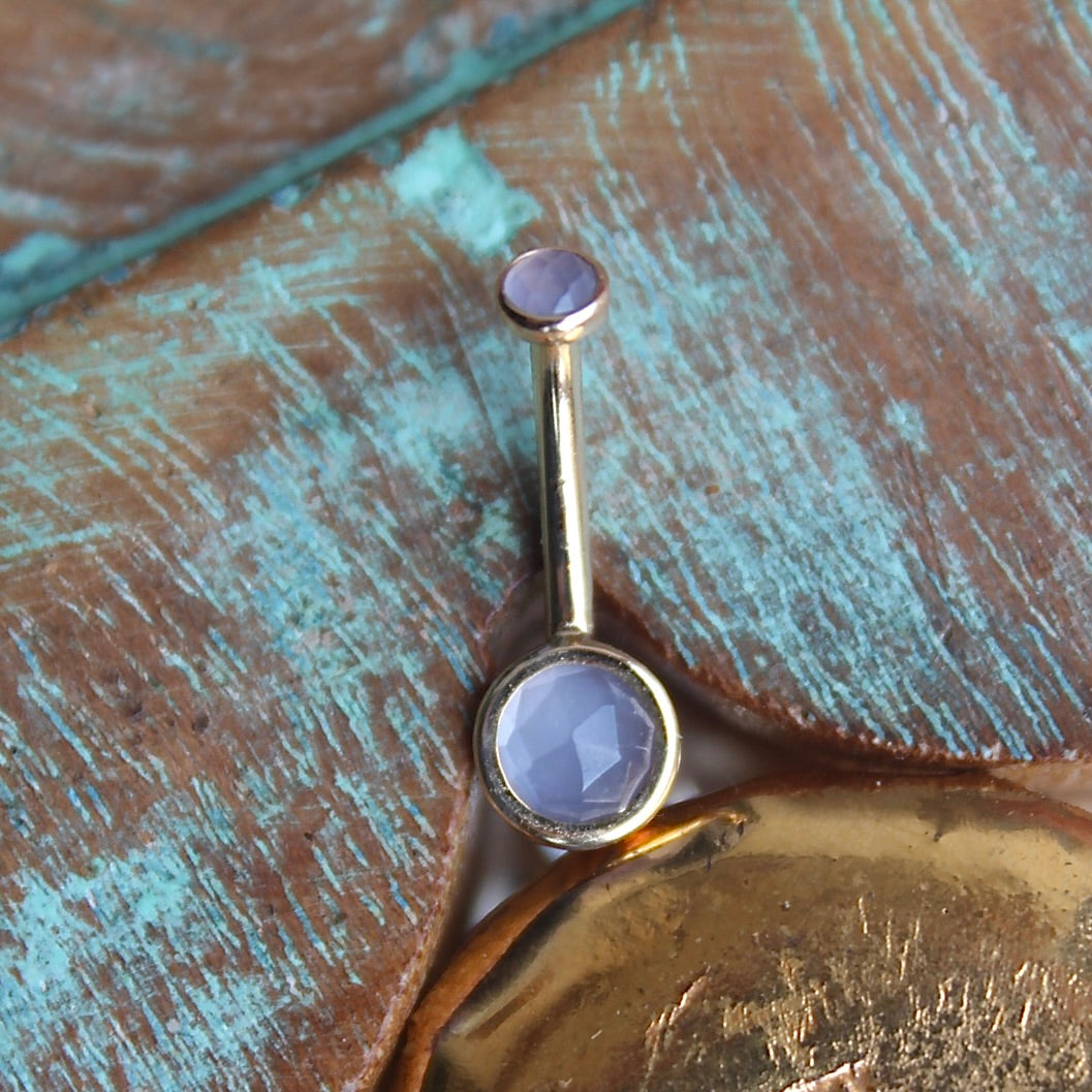 BVLA's "Classic Bezel Navel Curve" shown in 14k Yellow gold with lavender chalcedony. Shown on a background featuring a piece of wood with blue paint on top and a small golden piece on the bottom of the photo