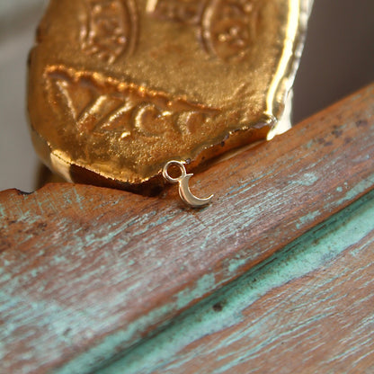 BVLA's "Flat Moon Charm" shown in 14k Yellow gold. Shown on a background featuring a piece of wood with blue paint on bottom and a small golden piece on the top of the photo