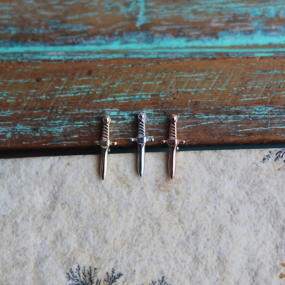 BVLA's "Slasher Dagger" shown from left to right in 14k Yellow gold, 14k White gold and 14k Rose gold. Shown on a background featuring a piece of wood with blue paint on top and a light grey stone on the bottom of the photo