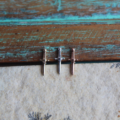 BVLA's "Slasher Dagger" shown from left to right in 14k Yellow gold, 14k White gold and 14k Rose gold. Shown on a background featuring a piece of wood with blue paint on top and a light grey stone on the bottom of the photo