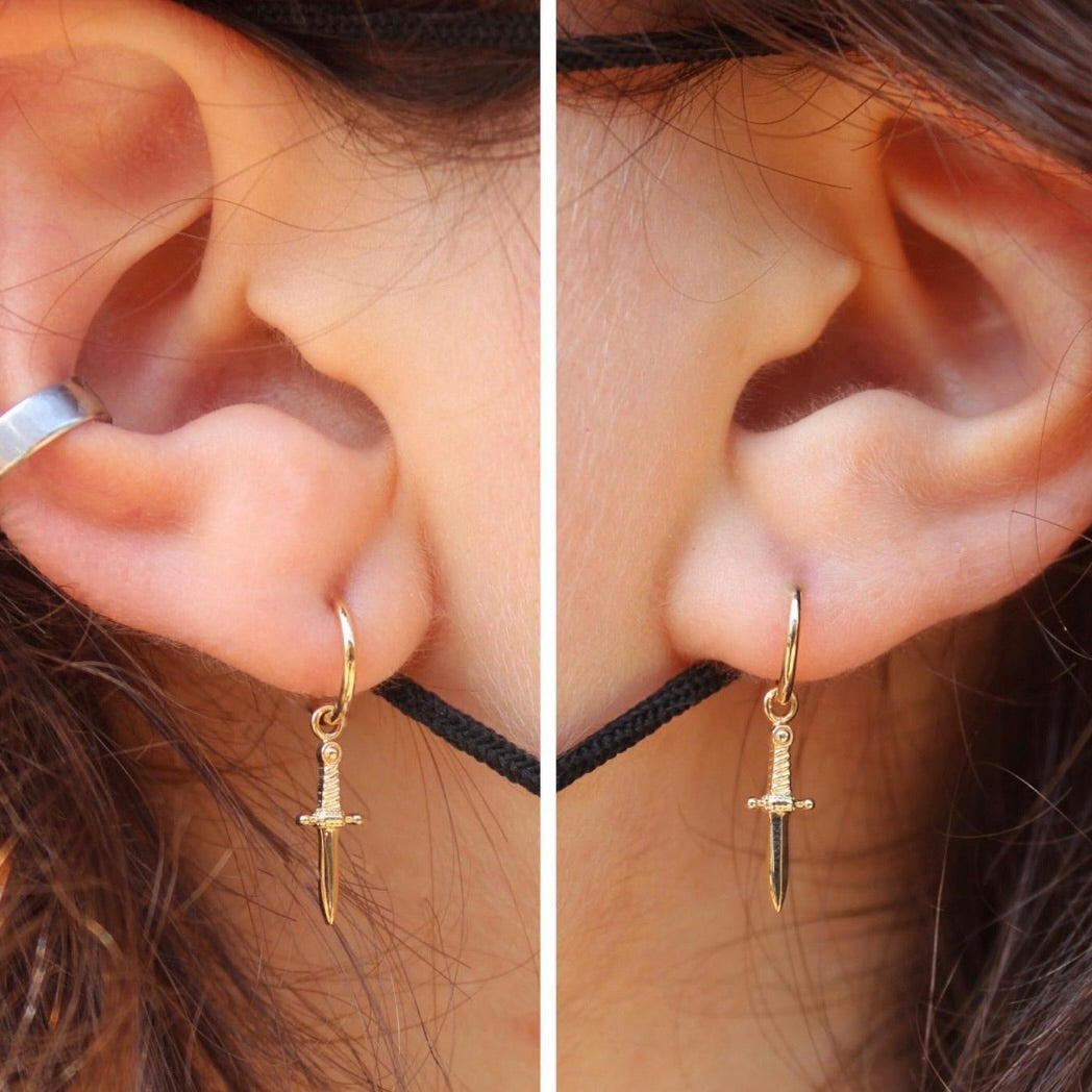 
                  
                    Left and right ears featuring a healed lobe piercings with fitted 14k Yellow Gold BVLA "Seam Ring" with a 14k Yellow Gold BVLA "Slasher Dagger Charm" dangling from them
                  
                