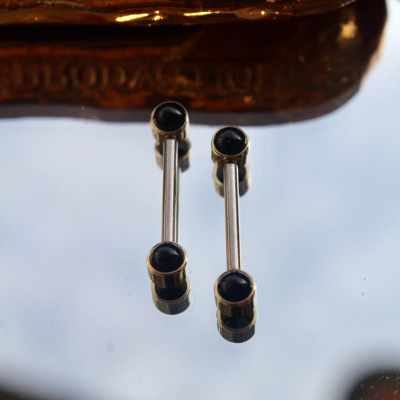 
                  
                    BVLA's "Bezel Side Pin" shown 4 times in 14k Yellow gold with onyx, 2 each attached to 2 Anatometal "Straight Barbell" in titanium. Shown on a background featuring a mirror and a small golden piece on the top
                  
                