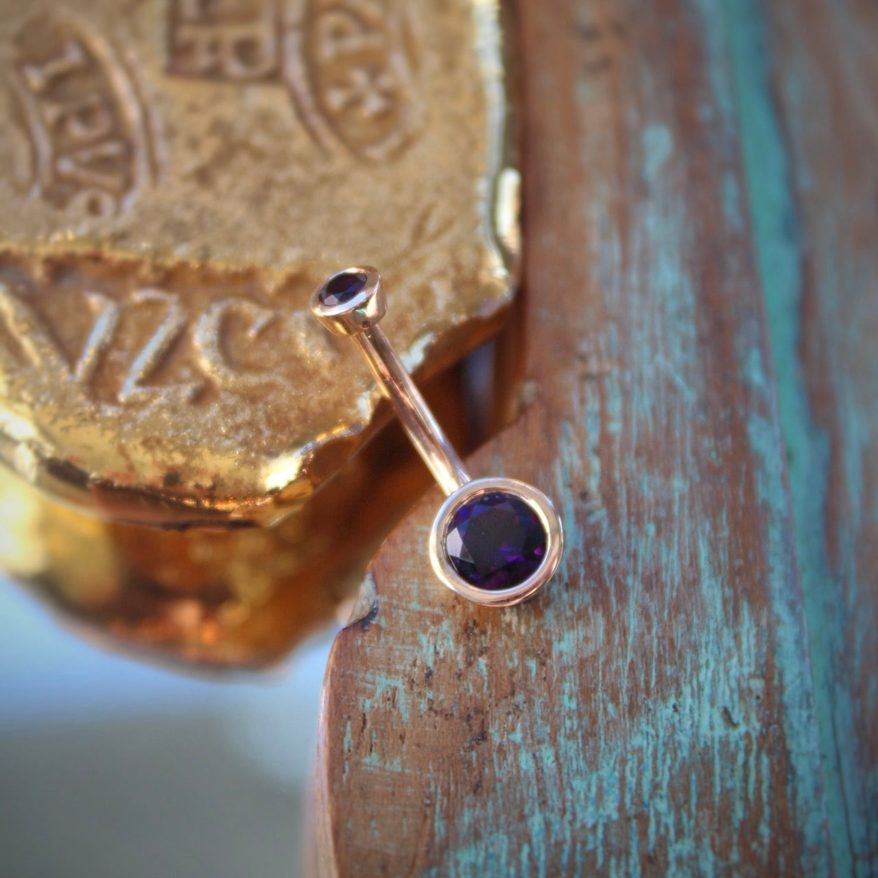 BVLA's "Classic Bezel Navel Curve" shown in 14k Rose gold with amethyst. Shown on a background featuring a piece of wood with blue paint on bottom right and a small golden piece on the top left of the photo