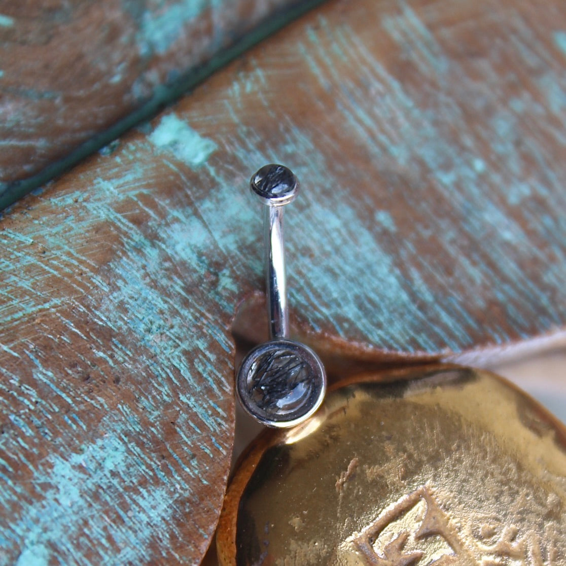 BVLA's "Classic Bezel Navel Curve" shown in 14k White gold with tourmalated quartz. Shown on a background featuring a piece of wood with blue paint on top and a small golden piece on the bottom of the photo