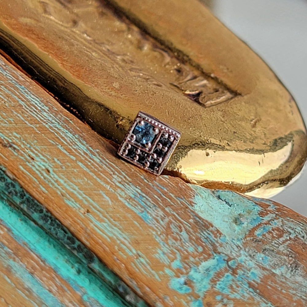 
                  
                    BVLA's "Endymoin Square" shown in 14k White gold with 1 Swiss blue topaz and 5 smaller London blue topaz. Shown on a background featuring a piece of wood with blue paint on bottom and a small golden piece on the top right of the photo
                  
                