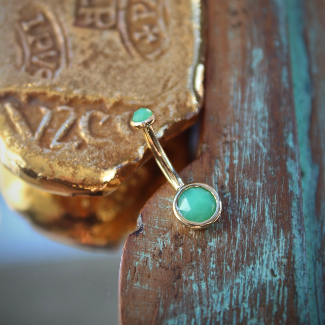 BVLA's "Classic Bezel Navel Curve" shown in 14k Yellow gold with chrysoprase. Shown on a background featuring a piece of wood with blue paint on bottom right and a small golden piece on the top left of the photo