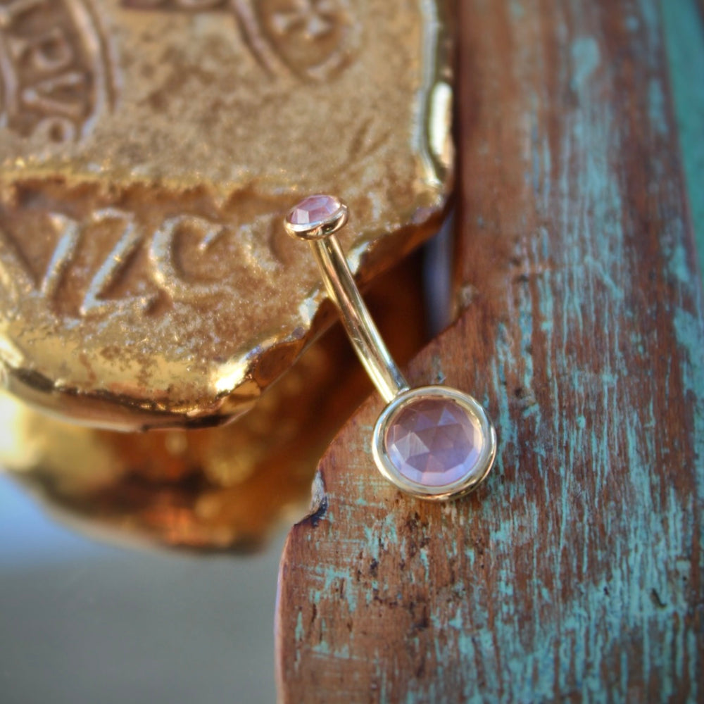 
                  
                    BVLA's "Classic Bezel Navel Curve" shown in 14k Yellow gold with rose quartz. Shown on a background featuring a piece of wood with blue paint on bottom right and a small golden piece on the top left of the photo
                  
                