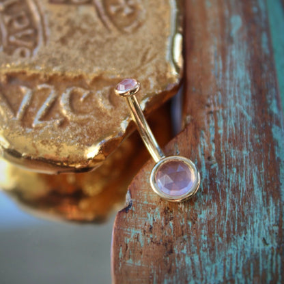 BVLA's "Classic Bezel Navel Curve" shown in 14k Yellow gold with rose quartz. Shown on a background featuring a piece of wood with blue paint on bottom right and a small golden piece on the top left of the photo