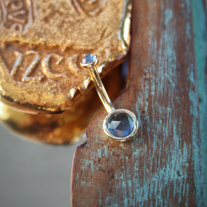 BVLA's "Classic Bezel Navel Curve" shown in 14k Yellow gold with swiss blue topaz. Shown on a background featuring a piece of wood with blue paint on bottom right and a small golden piece on the top left of the photo