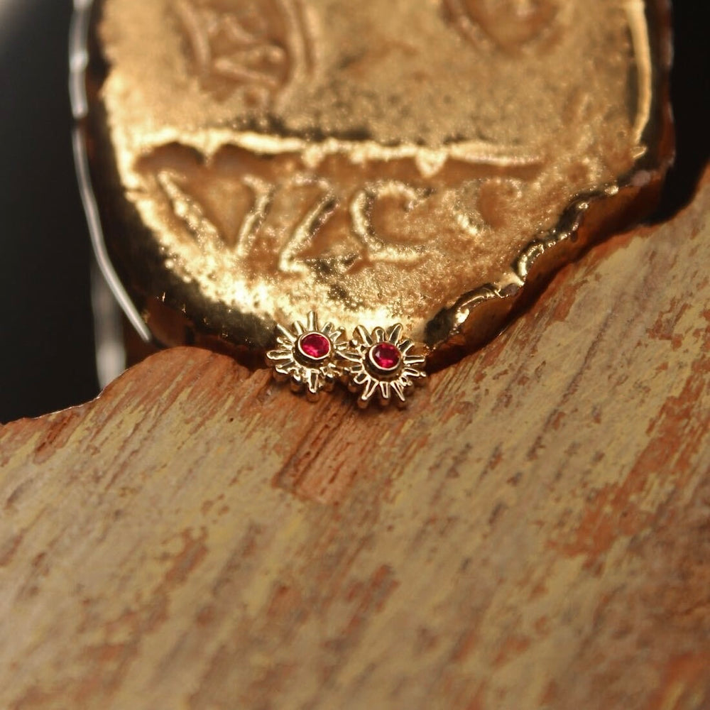 
                  
                    BVLA's "Sun Ray" shown twice in 14k Yellow gold with ruby. Shown on a background featuring a piece of wood with blue paint on bottom and a small golden piece on the top of the photo
                  
                