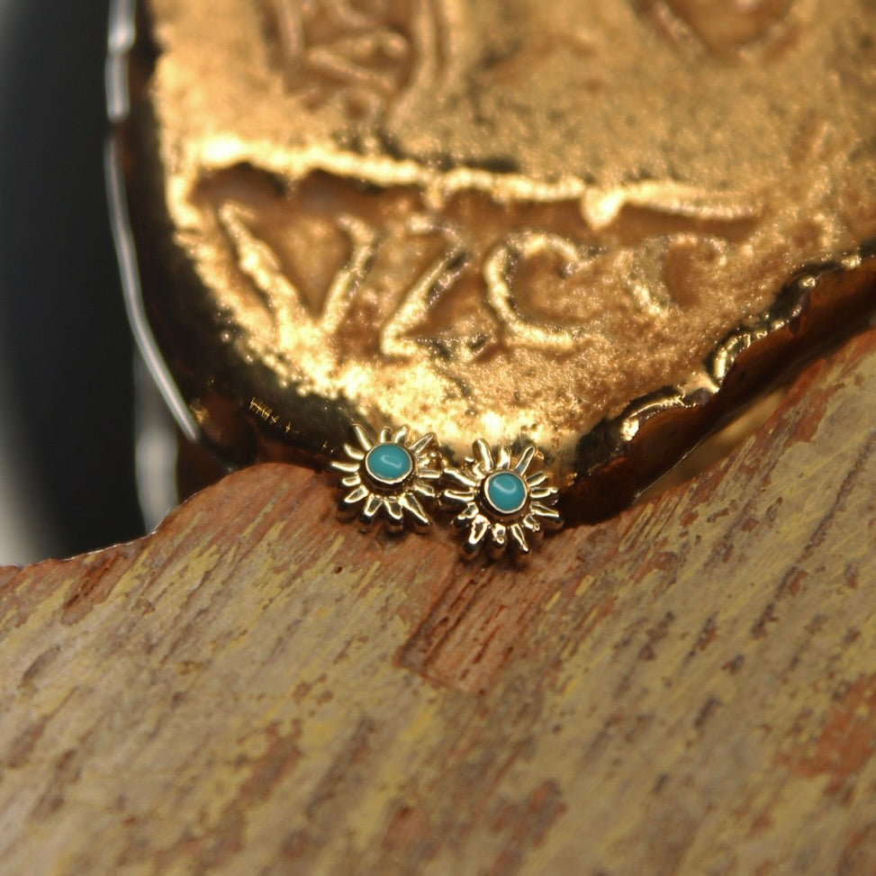 
                  
                    BVLA's "Sun Ray" shown twice in 14k Yellow gold with turquoise. Shown on a background featuring a piece of wood with blue paint on bottom and a small golden piece on the top of the photo
                  
                
